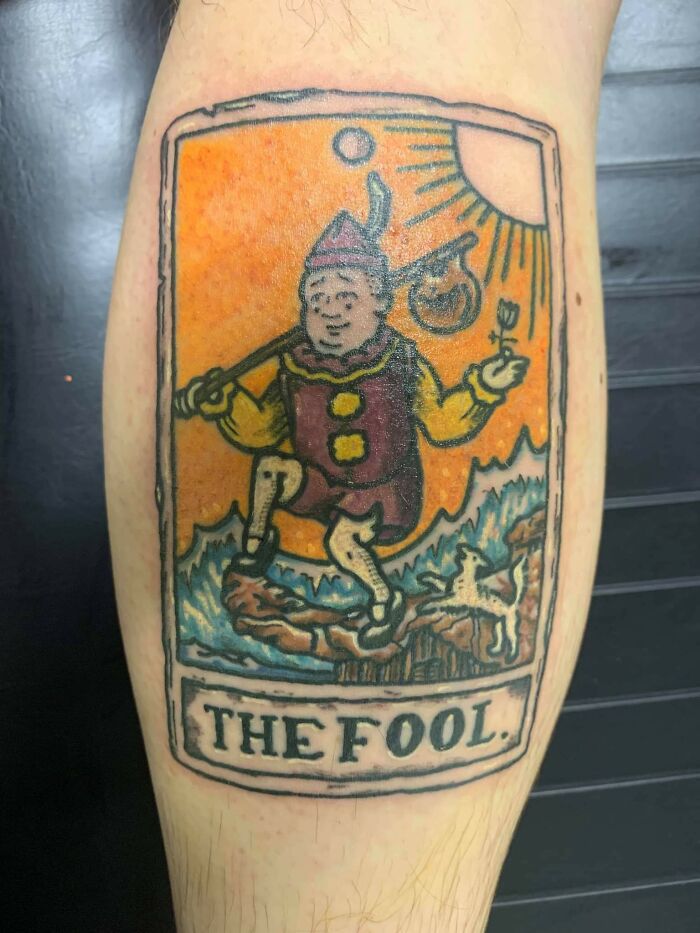Colorful tarot card tattoo of The Fool on a leg, a cool tattoo design with bright orange and blue tones.