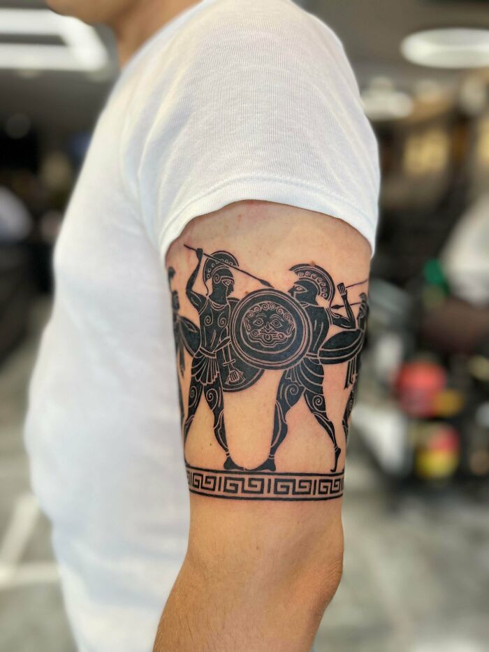 Tattoo of ancient Greek warriors with shields and spears in black ink on a person's upper arm, cool tattoo design.