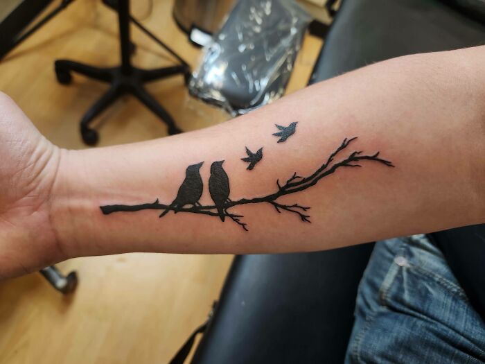 Black tattoo of birds sitting on a branch on a forearm, showcasing one of the cool tattoos without regrets.