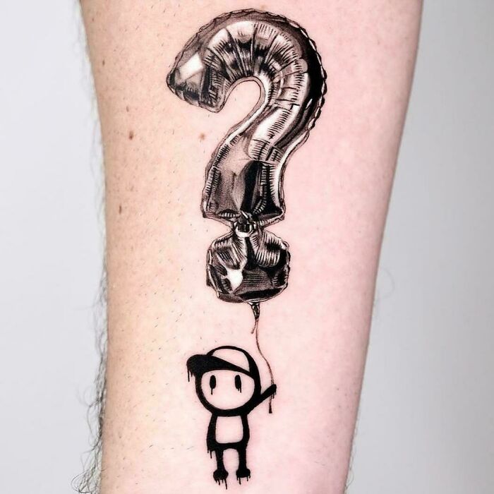 Black ink tattoo of a small cartoon figure holding a balloon shaped like a question mark on forearm, cool tattoo design.