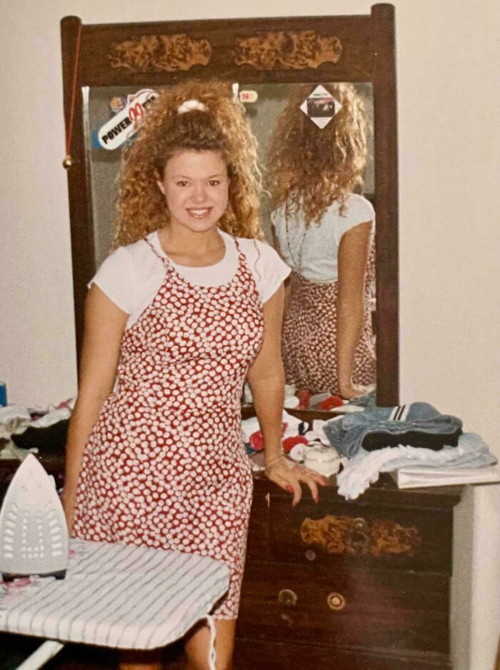 Young woman with curly hair smiling in a vintage bedroom, standing near ironing board and dresser, evoking nostalgic Gen X vibes.