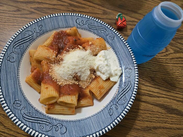 Plate of rigatoni pasta with tomato sauce and cheese, a nostalgic Gen X meal on a wooden table with a blue water bottle.