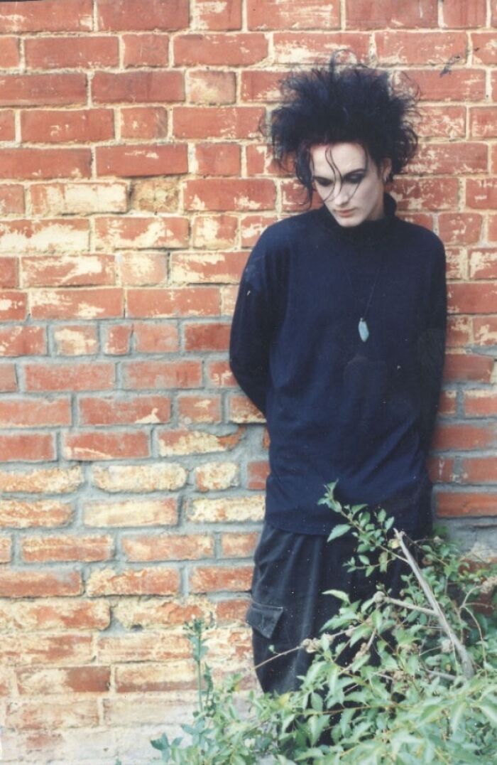 Young adult with messy hair and dark clothing leaning against a brick wall, capturing nostalgic Gen X vibes.