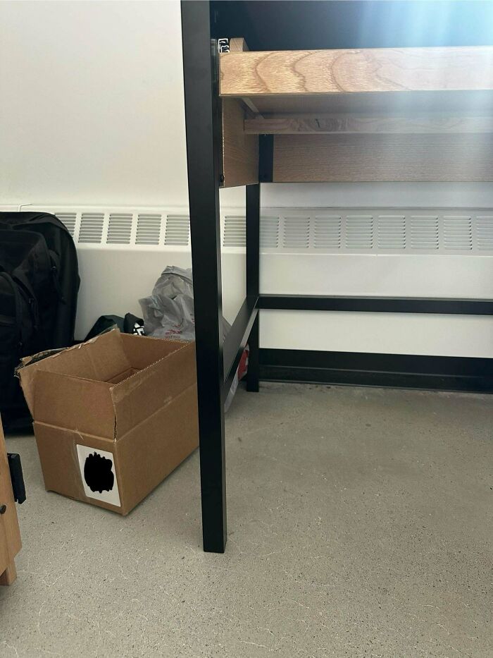 Cardboard box camouflaged on a gray floor next to black bed frame legs and a white wall radiator vent.