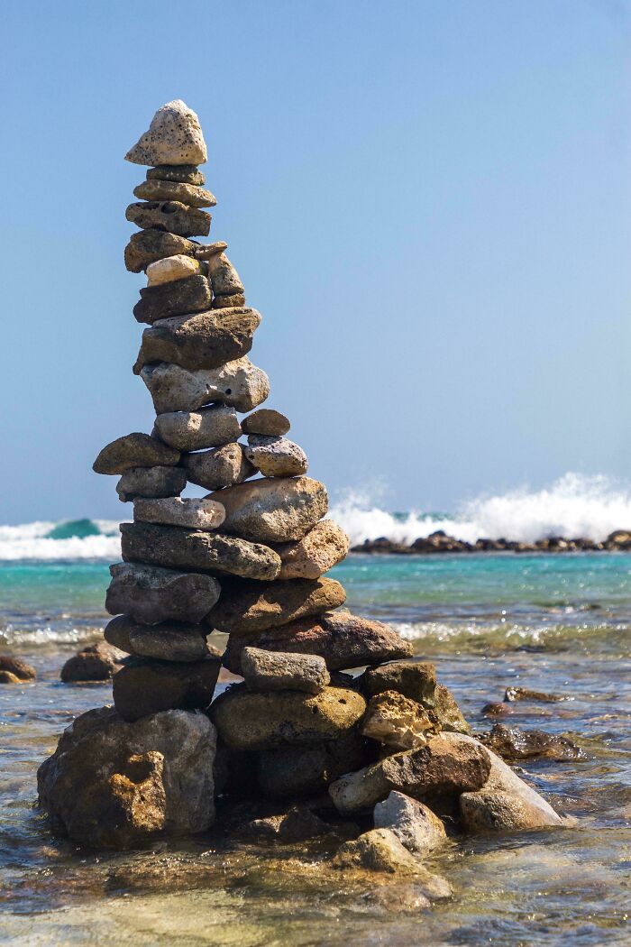 Stack of perfectly camouflaged stones balanced on a rocky shore with ocean waves in the background on a clear day