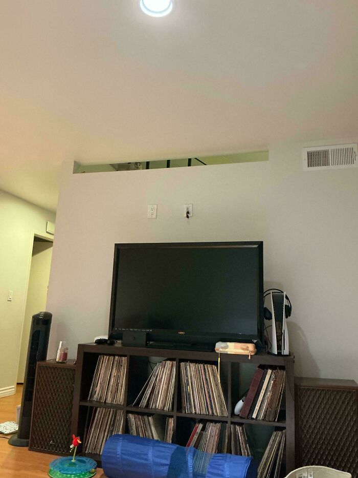 Cat perfectly camouflaged on a high ledge above a television in a living room, blending into the surroundings.