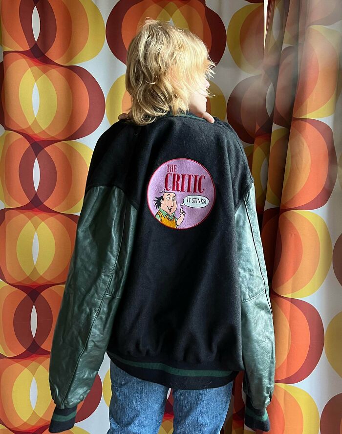 Person wearing a vintage 90s jacket with a cartoon patch against a retro orange and yellow patterned background.
