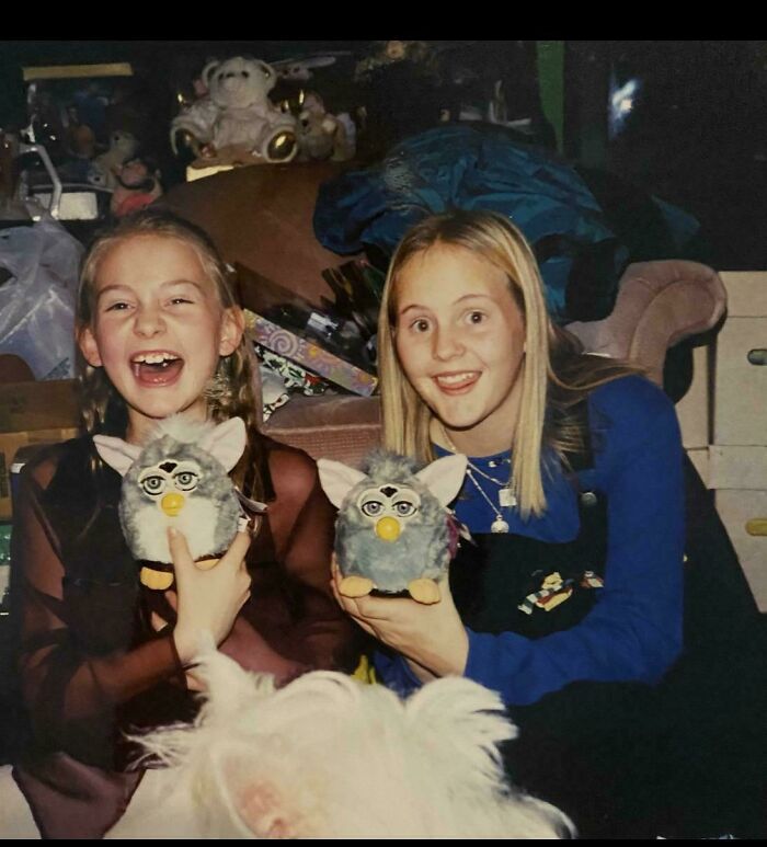 Two girls smiling and holding vintage Furby toys in a nostalgic 90s childhood setting filled with stuffed animals.
