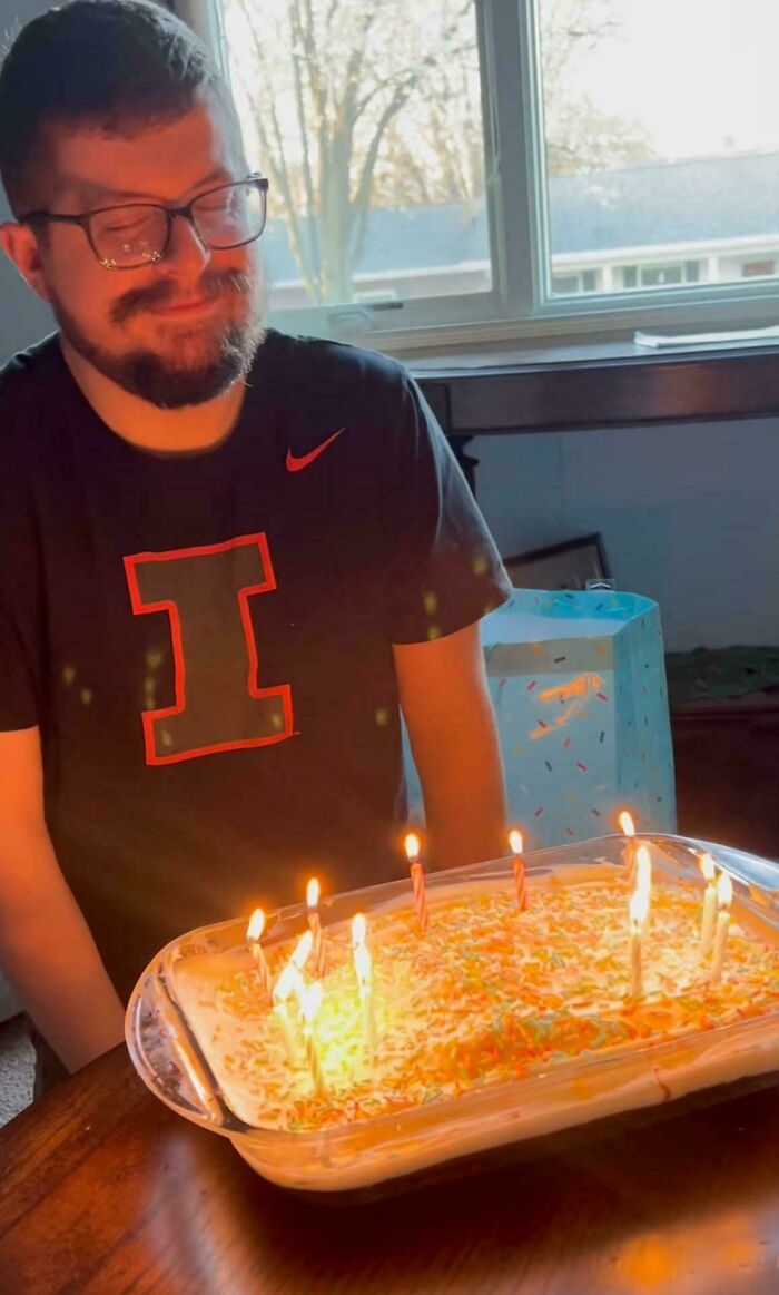 Man smiling at birthday cake with lit candles near a window