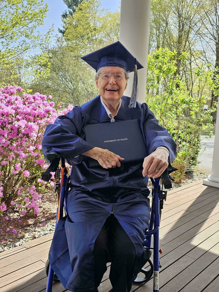 Elderly person smiling in graduation gown holding diploma outdoors
