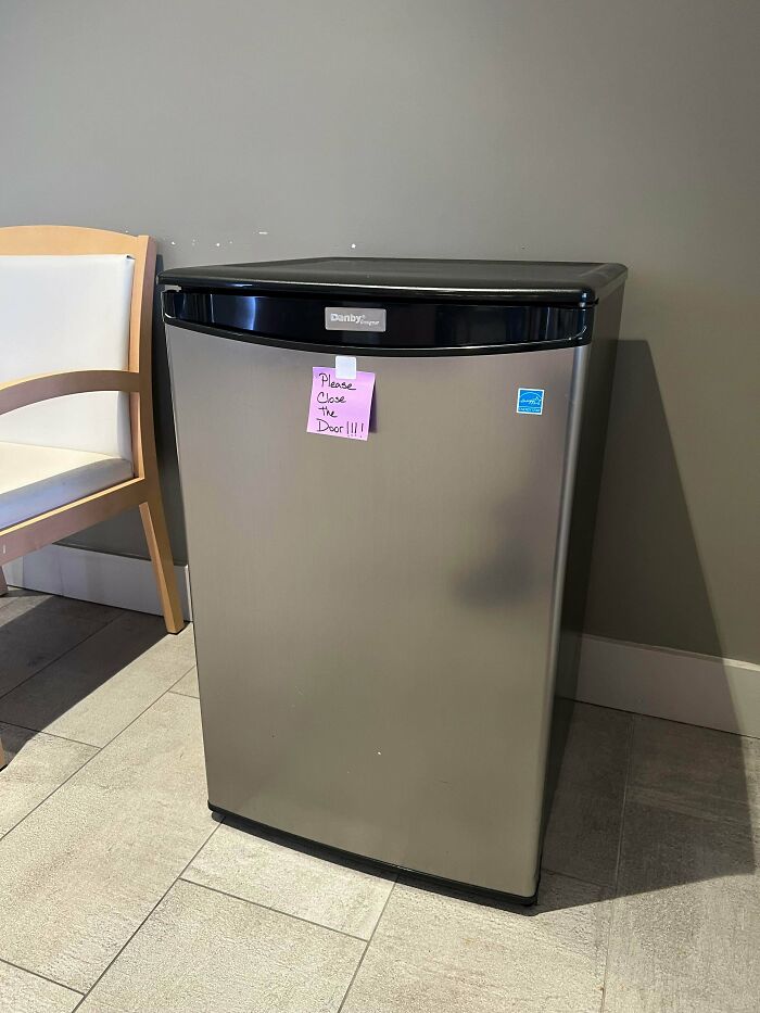 A small silver refrigerator with a passive-aggressive note on the door saying Please Close The Door!!!