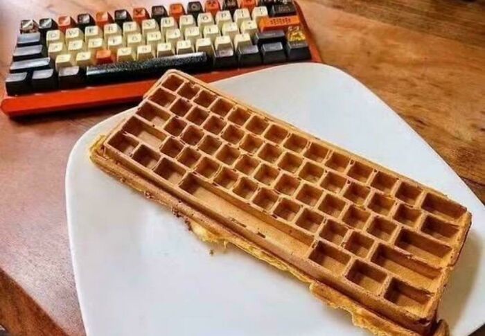 Waffle shaped like a keyboard on a plate beside a vintage mechanical keyboard in a wooden setting, weird confusing image.