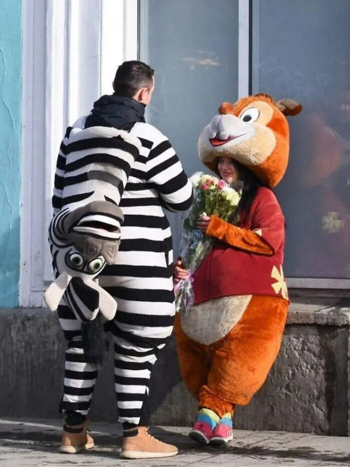 Two people in confusing costumes, one in a zebra suit with an upside-down animal, the other in a large chipmunk outfit holding flowers.