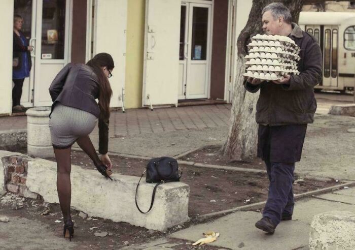 Woman bending over to tie shoe on a city sidewalk while man carries multiple trays of eggs in a confusing street scene.