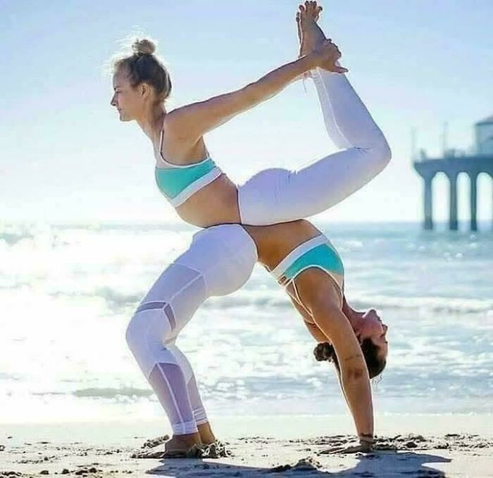 Two women performing a weird and confusing acrobatic yoga pose on the beach with ocean and pier background.