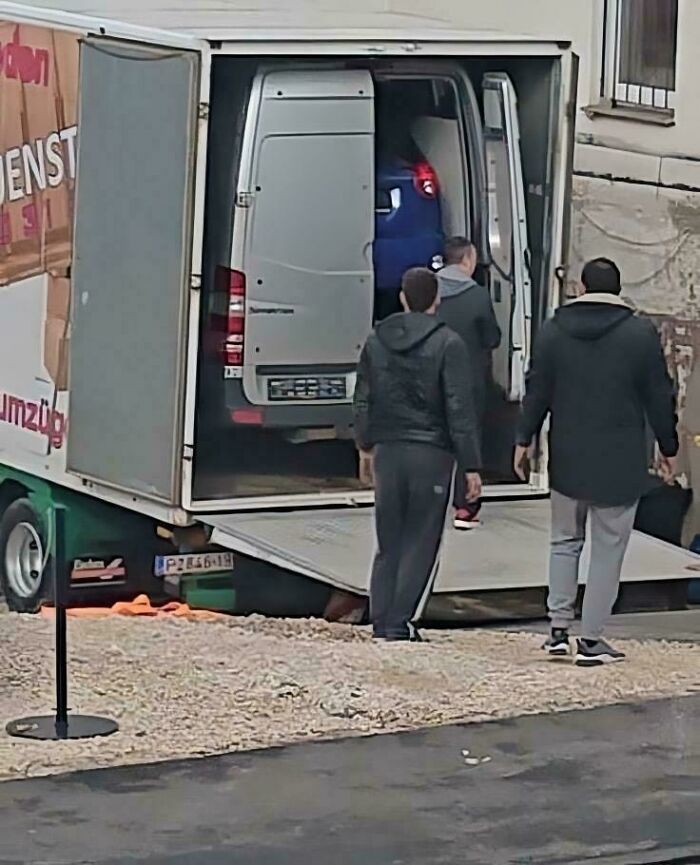 Unusual image showing a van inside a larger truck with people standing nearby, a weird and confusing scene.
