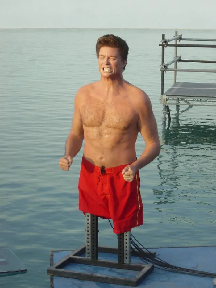 Shirtless man in red shorts with mechanical legs standing on a platform over water in a weird, confusing, and cursed image.