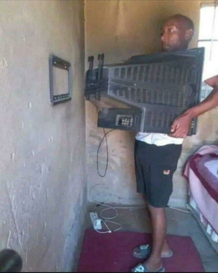 Man holding a large TV to his chest while facing a tiny wall-mounted screen in a weird and confusing cursed image.