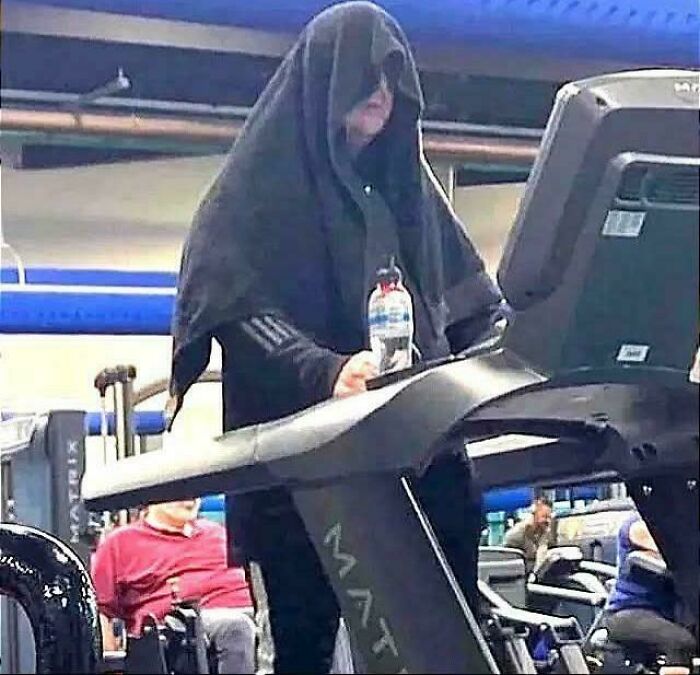 Person wearing a black hooded cloth over their head while walking on a treadmill in a gym, creating a weird, confusing scene