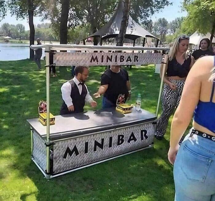 Mini bar set up outdoors with a man in a vest serving drinks, surrounded by people in a park setting on a sunny day.