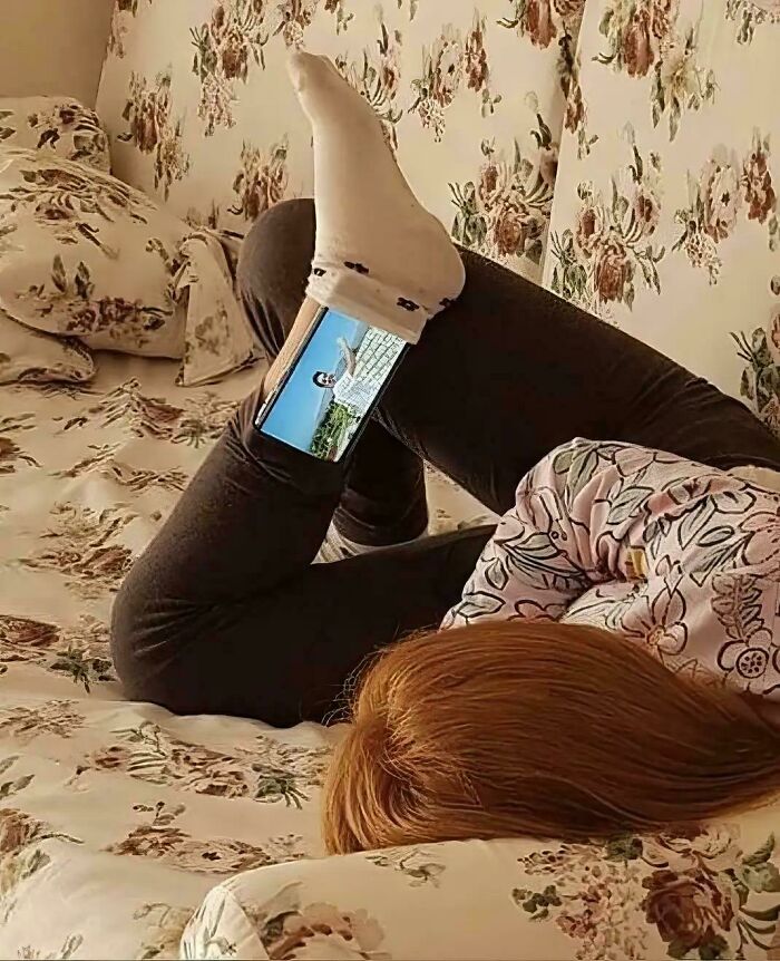 Person lying on floral bed with phone tucked behind leg showing video, a weird and confusing image.
