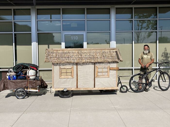 Tiny house on wheels with thatched roof hitched to bicycle parked outside building