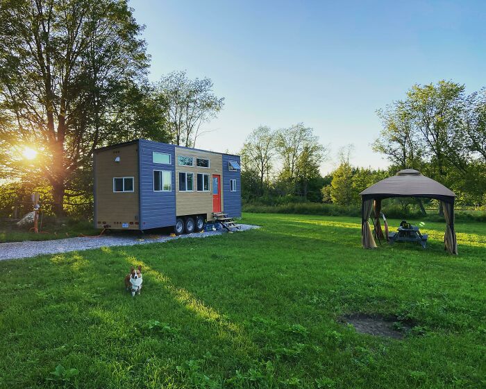 Small cozy tiny home on wheels in grassy yard with dog and gazebo nearby