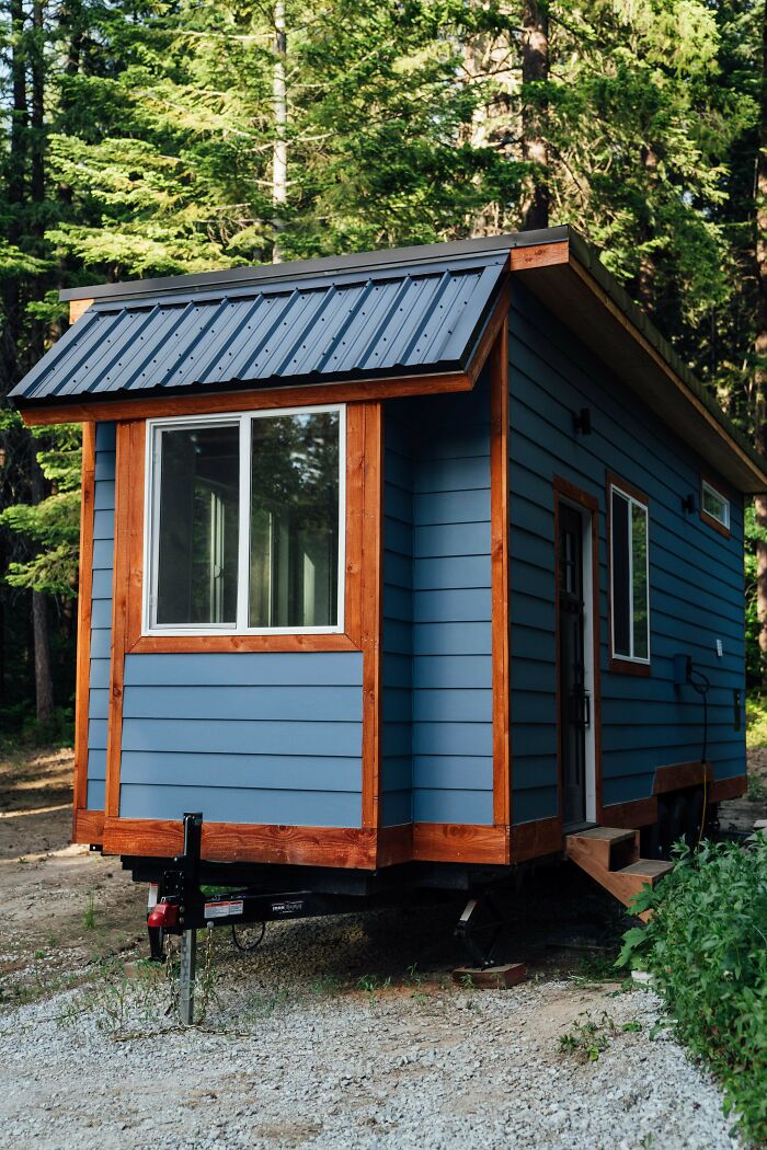 Blue tiny house with wood trim and metal roof parked in forest clearing