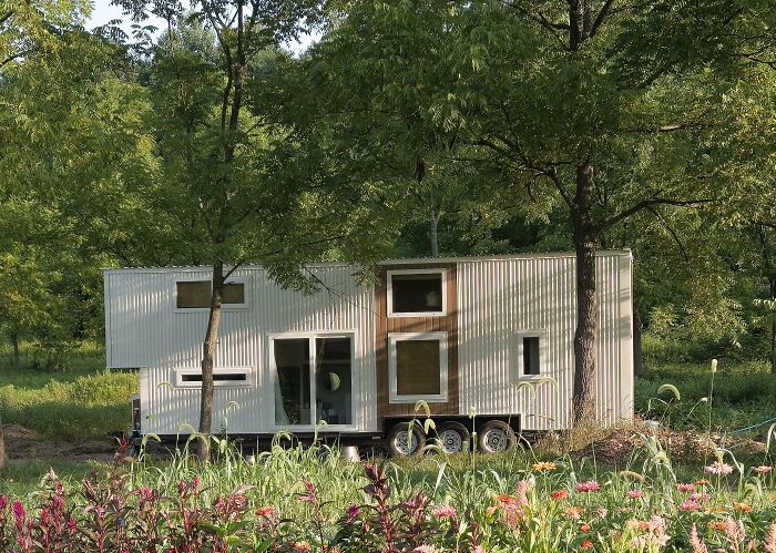 Modern tiny home on wheels surrounded by trees and flowers in a cozy natural setting