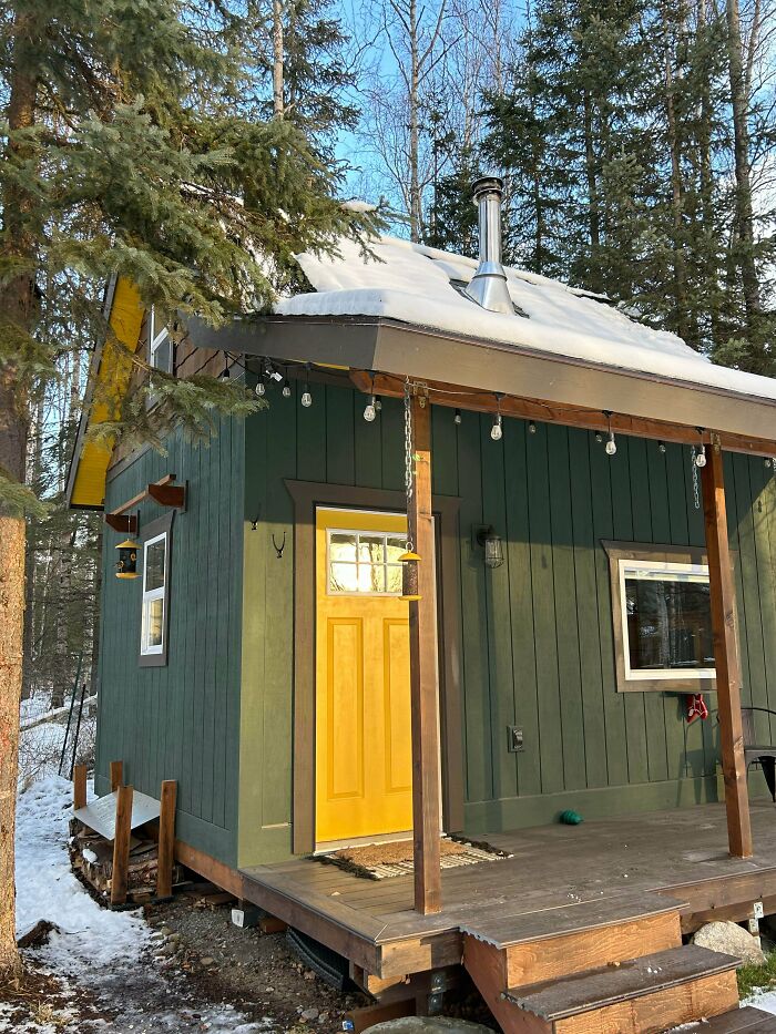 Green tiny house with yellow door and snow on roof in forest