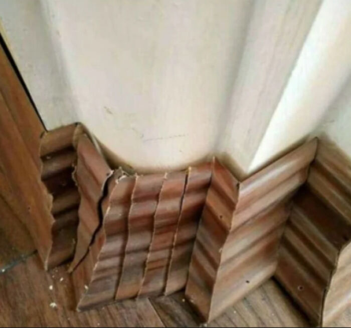 Warped wooden baseboard awkwardly installed in a corner, showing a technically works but hilariously dumb solution to a problem.
