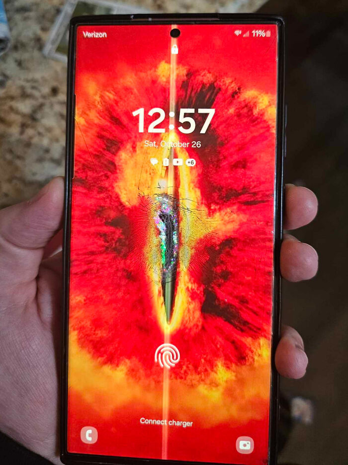 Smartphone with a cracked screen held in hand displaying a bright, fiery wallpaper and fingerprint unlock icon.