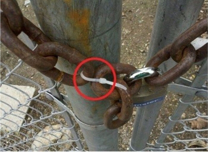 Rusty chain secured to metal gate post with a zip tie in a technically works but hilariously dumb solution.
