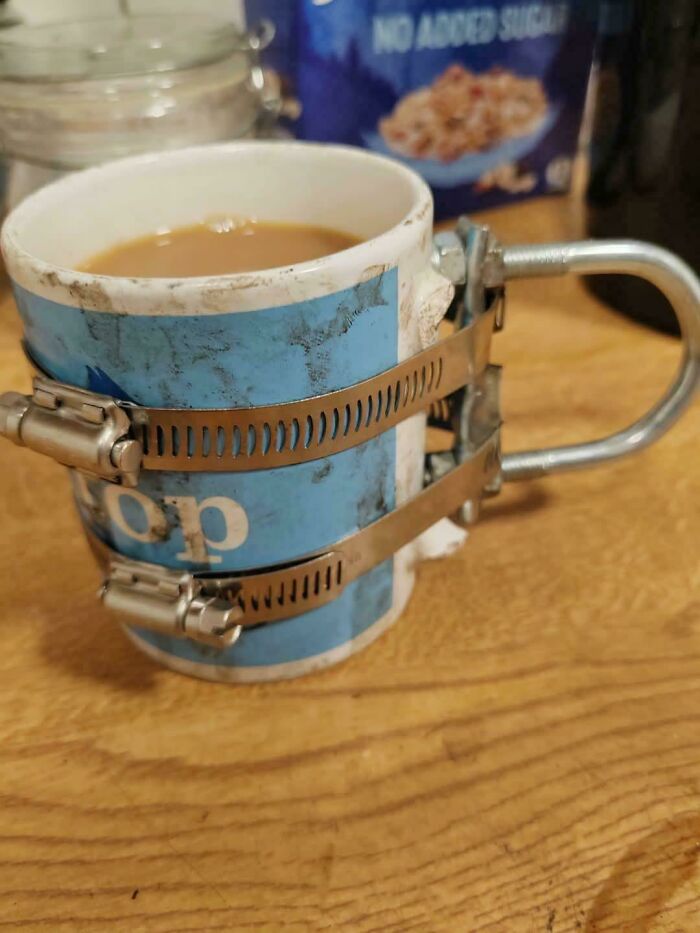 Coffee mug with broken handle fixed using metal hose clamps and a bent metal piece, a technically works DIY solution.