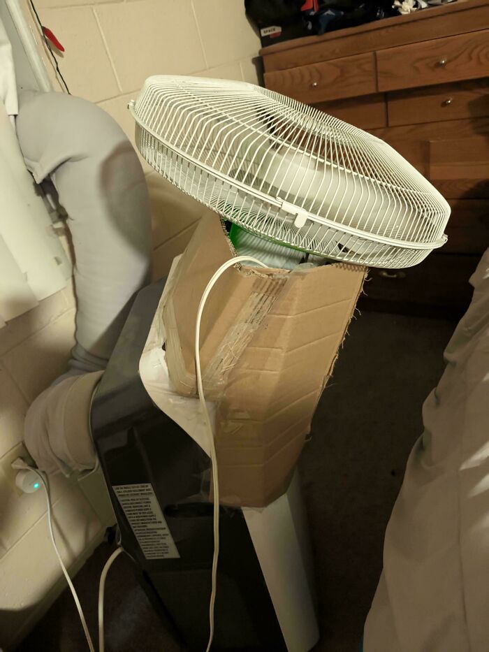 Improvised cooling setup with a fan taped to a cardboard attached to an air conditioner showing a technically works solution.