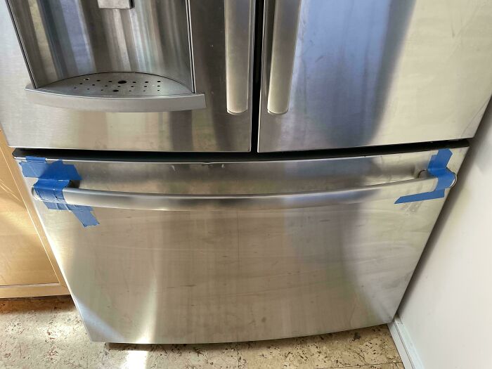 Stainless steel refrigerator drawer held shut with blue tape as a technically works DIY solution to a problem.