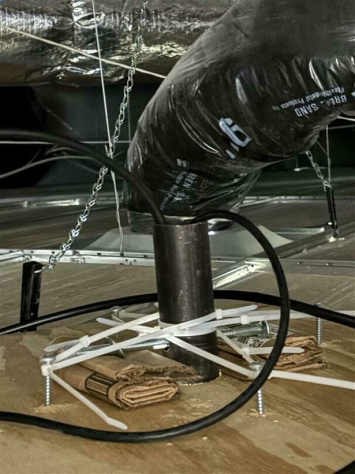 Improvised tech repair using zip ties, screws, and cardboard to secure a pipe in an awkward setup under a floor.