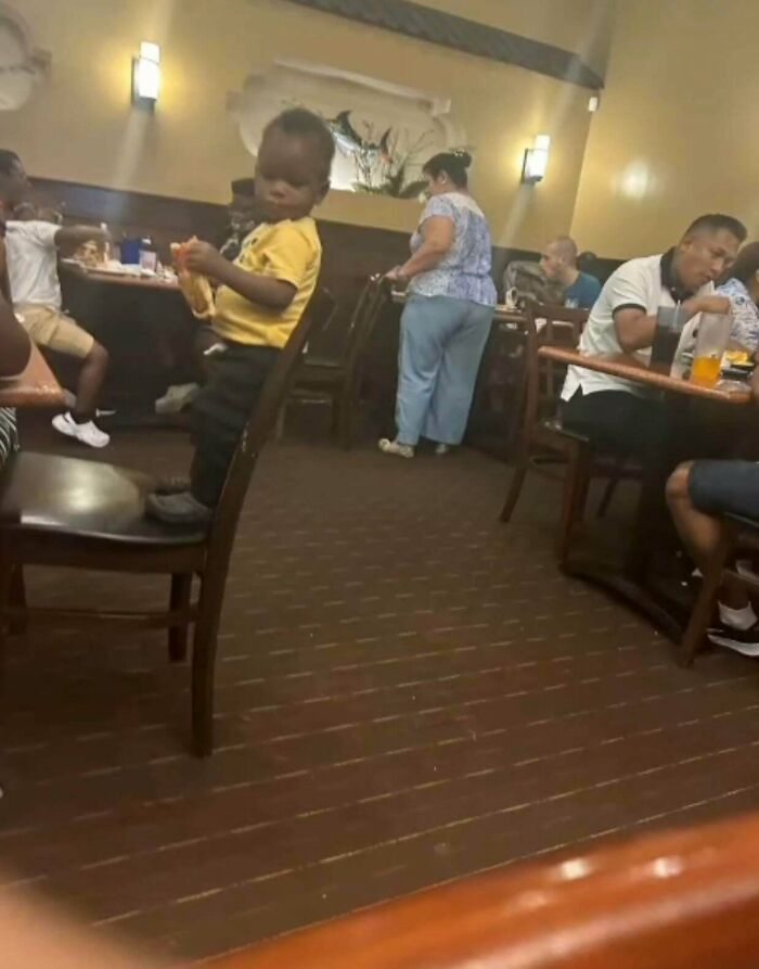 A kid in a yellow shirt stands on a chair at a restaurant, holding food. Dumb and funny kid moments.