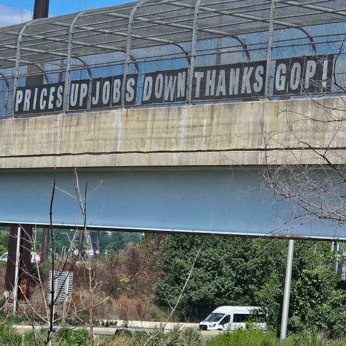 Concrete pedestrian bridge with a confusing protest sign reading prices up jobs down thanks gop under blue sky.
