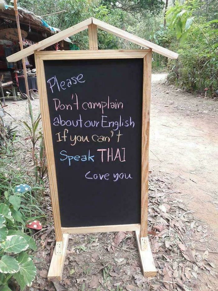 Chalkboard sign with a humorous message about language, outdoors on a dirt path surrounded by plants and trees.