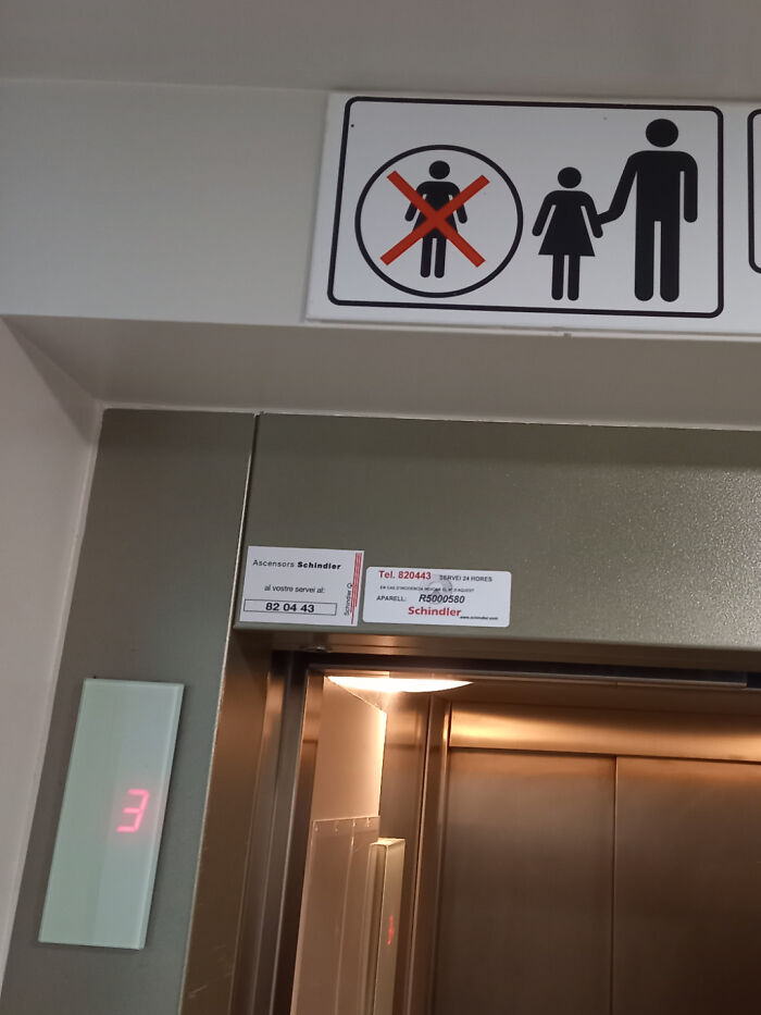 Elevator with a funny and confusing sign showing a man holding a child’s hand while a crossed-out child figure appears nearby.