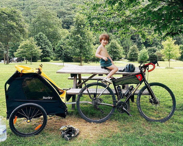 A happy child sits on a picnic table next to a bike with a trailer, celebrating dads and outdoor fun.