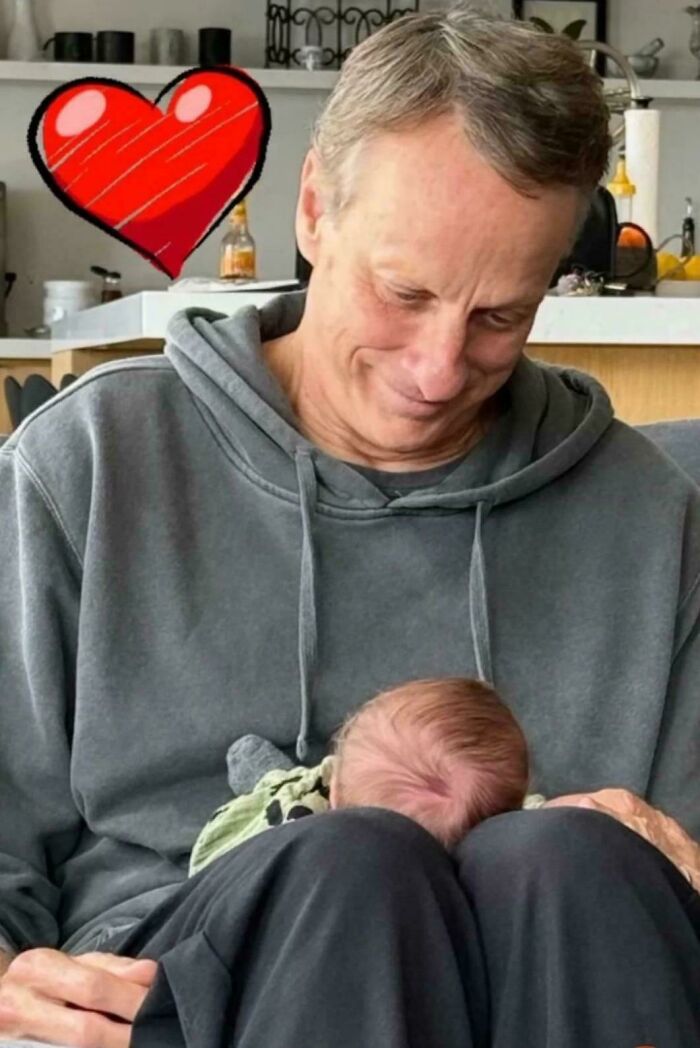 A gray-haired man in a hoodie smiles down at a baby sleeping on his lap. A red heart hovers above, celebrating Dads.