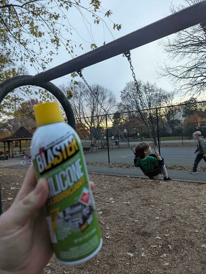 A hand holds a can of Blaster Silicone Lubricant at a playground with a child on a swing. Dads community.