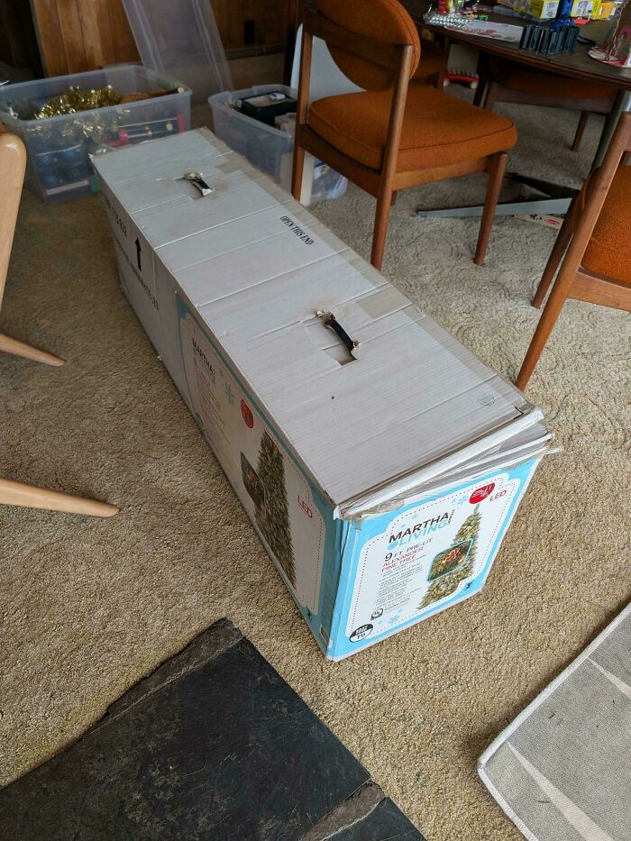 A new artificial Christmas tree box on a carpeted floor, with other boxes and furniture in the background. Good for dads.
