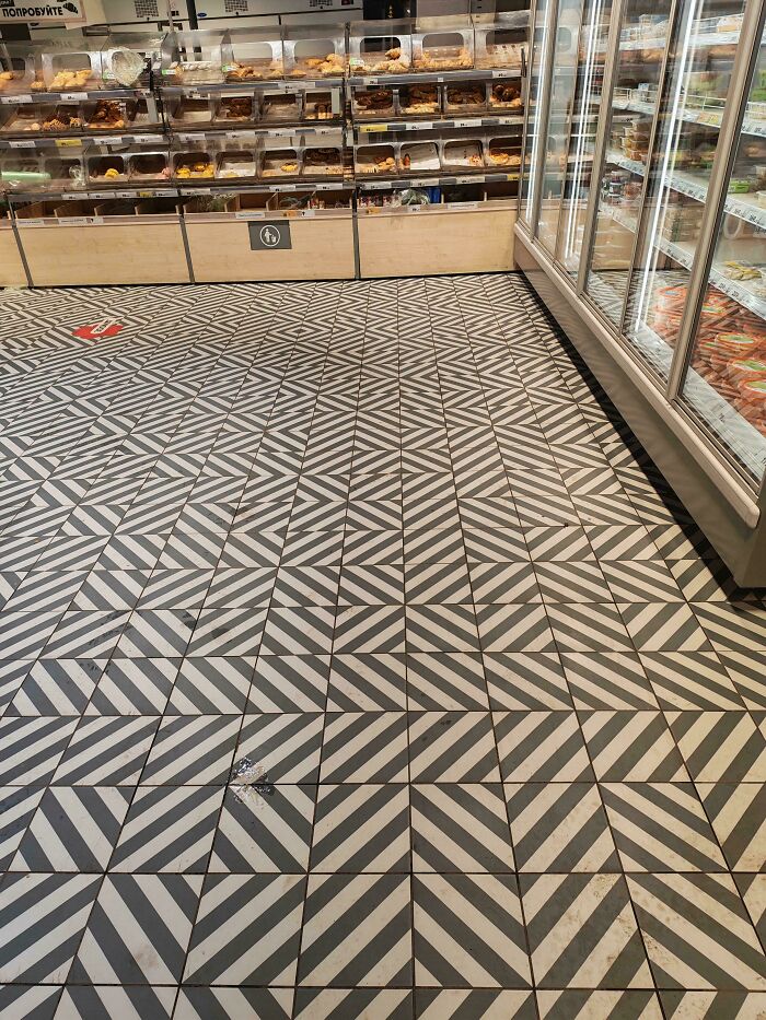 Geometric patterned floor tiles create a visually confusing design in a store with refrigerated and baked goods sections.
