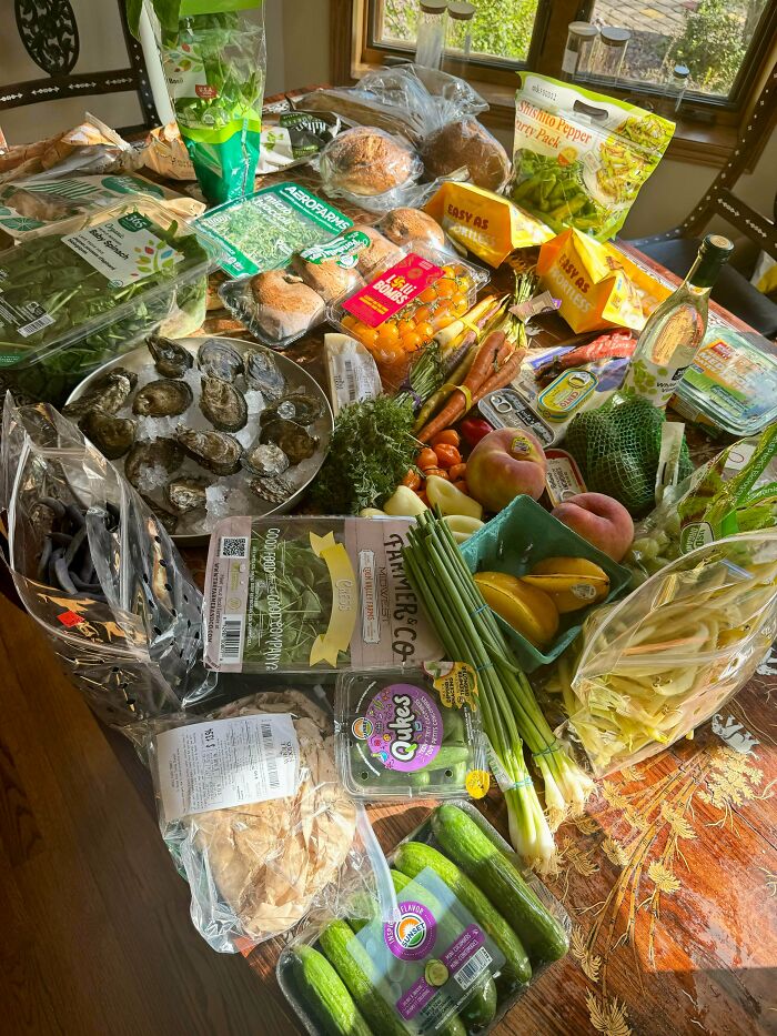 Variety of fresh groceries including vegetables, fruits, bread, and seafood displayed on a wooden table showing local grocery costs.