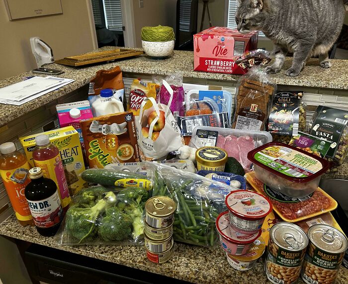 Assorted groceries including produce, canned goods, dairy, and snacks displayed on a counter showing grocery cost impact.