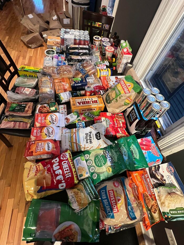 A large assortment of groceries including snacks, meats, drinks, and packaged foods displayed on a wooden table showing grocery costs.