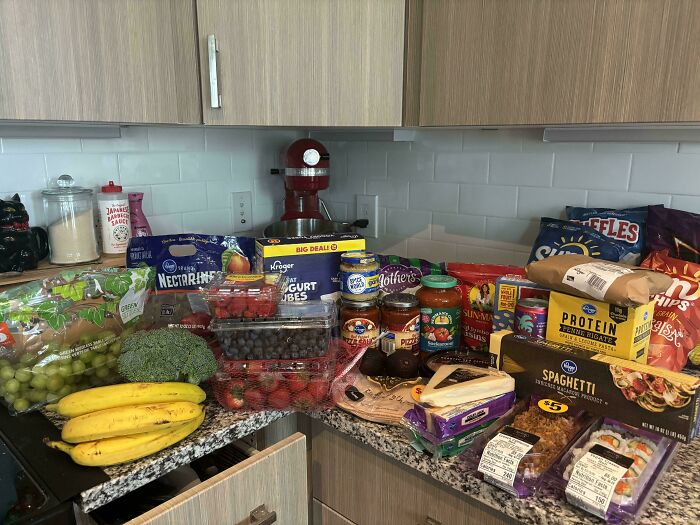 Groceries including fruits, vegetables, dairy, pasta, chips, and sauces displayed on a kitchen counter showing local grocery costs.